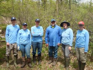 Six people posing for a picture in the woods. All wear matching work shirts, hats, and work boots.