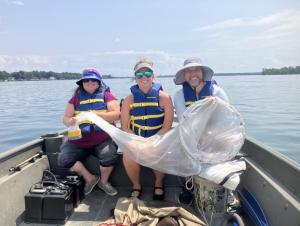 Three people smile and sit on the deck of a small boat on a sunny day, holding a conical net across all of their laps