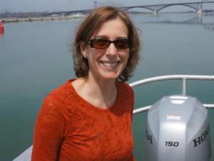 A smiling woman with sunglasses and shoulder length hair poses for a picture on the back of a boat in a canal