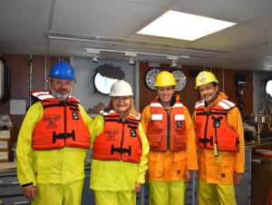 Four people wearing rain gear, hard hats, and life jackets indoors on a boat.