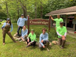 Seven people pose for a picture around a sign "Craneridge, established 1967." There are trees behind them and a shelter with mailboxes nearby.