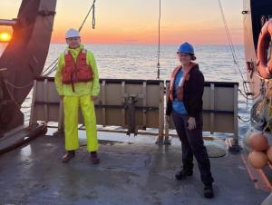 Two people wearing hard hats and life jackets stand on the back deck of a large boat on a lake at sunrise