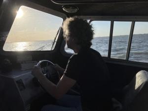 A person sits at the helm inside the cabin of a boat. The sun shines brightly through the window, casting everything inside into shade