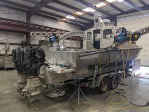 A metal boat with a cabin sits on a trailer in a large garage. The back of the boat is under construction, and the two large motors are uncovered.