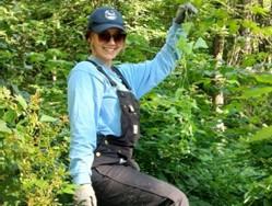A light-skinned person stands in the brush of a forest wearing a hat, sunglasses, overalls, and work gloves