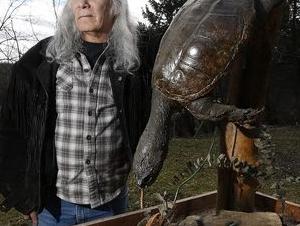 a man with long grey hair stands next to a taxidermied turtle near some trees