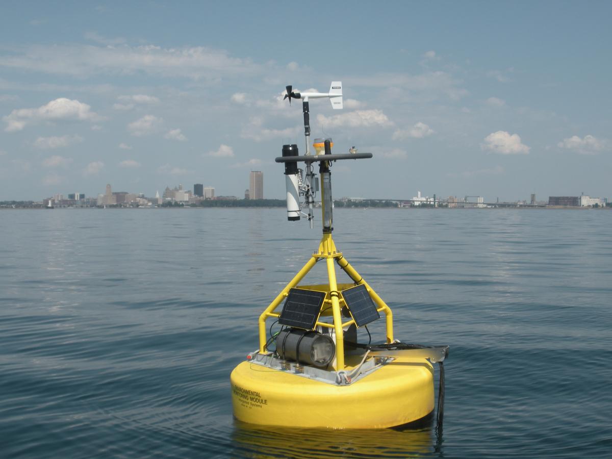 A buoy with meteorological equipment and solar panels floats in a big body of water near a city.