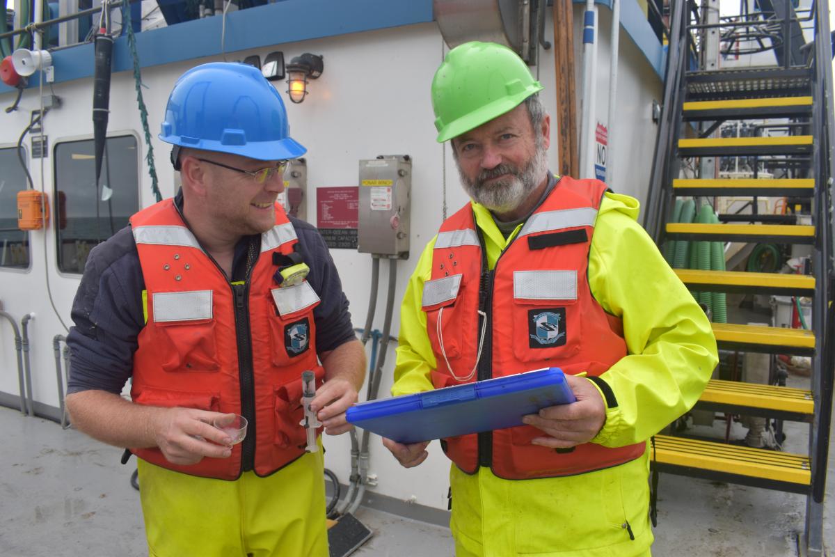 Two people wearing hard hats and life jackets stand on a large boat near a staircase. One is holding a clipboard and the other has a syringe.